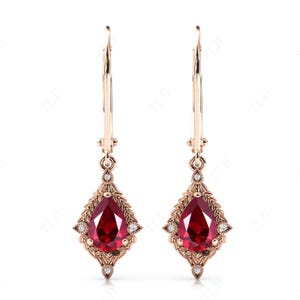 May include: A pair of rose gold drop earrings. Each earring features a pear-shaped, deep red gemstone set within a diamond-shaped frame with intricate detailing. Small, clear gemstones accent the frame. The earrings hang from a lever back closure.