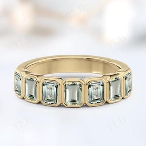 Può includere: Un anello a fascia in oro con una serie di pietre preziose rettangolari verde chiaro. L'anello ha una finitura dorata liscia e lucida e le pietre preziose sono incastonate in castoni individuali. È un gioiello.