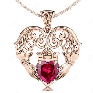 May include: A gold-tone pendant necklace featuring a heart-shaped ruby gemstone surrounded by two clasped hands, a traditional Irish symbol known as the Claddagh. The pendant is adorned with intricate filigree details.