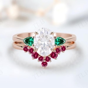 Puede incluir: Anillo de oro rosa con una piedra blanca de talla ovalada, flanqueada por dos piedras verdes en forma de pera y una hilera de pequeñas piedras rojas. El anillo tiene un diseño de doble banda.