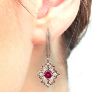 May include: A pair of dangle earrings with a red gemstone surrounded by smaller white gemstones. The earrings are made of gold and have a delicate, intricate design.