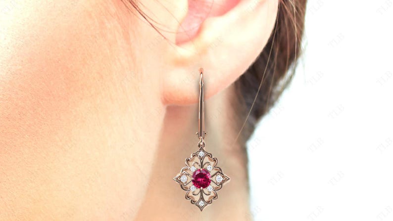 May include: A pair of dangle earrings with a red gemstone surrounded by smaller white gemstones. The earrings are made of gold and have a delicate, intricate design.