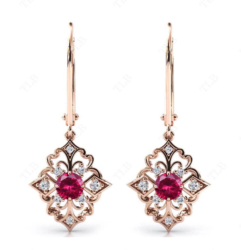 May include: A pair of rose gold dangle earrings with a delicate filigree design. Each earring features a round red ruby gemstone surrounded by smaller clear gemstones.