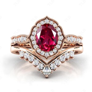 Könnte beinhalten: Ein Verlobungsring aus Roségold mit einem großen ovalen Rubin, der von einem Heiligenschein aus kleineren Diamanten umgeben ist. Der dazugehörige Ehering hat ein zartes V-förmiges Design mit Pavé-Diamanten.