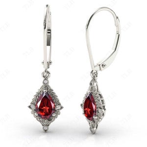 May include: A pair of silver dangle earrings with a pear-shaped red gemstone surrounded by smaller clear gemstones. The earrings have a leverback closure.