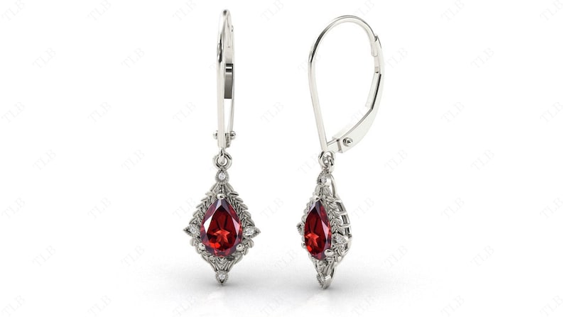 May include: A pair of silver dangle earrings with a pear-shaped red gemstone surrounded by smaller clear gemstones. The earrings have a leverback closure.