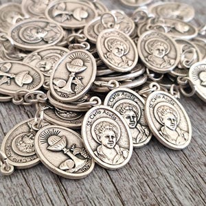 Puede incluir: Una colección de dijes ovalados de plata con imágenes religiosas. Los dijes presentan imágenes de un cáliz, un santo y el texto "St. Carlo Acutis".