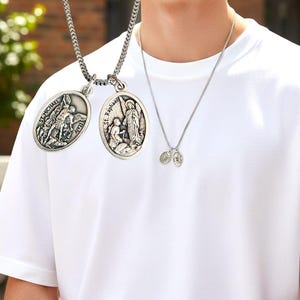 Op de afbeelding: Zilverkleurige hangerkettingen. Eén ketting heeft twee ovale hangers met religieuze afbeeldingen en de tekst "St. Michael" en "St. Raphael". Een andere ketting heeft twee kleinere ovale hangers. De kettingen worden gedragen over een wit T-shirt.