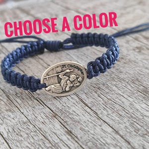 Könnte beinhalten: Ein marineblaues geflochtenes Armband mit einem silbernen St. Christopher-Medaillon. Das Armband hat verstellbare Gleitknoten und den Text "CHOOSE A COLOR" in Pink.