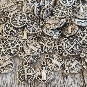 Puede incluir: Una colección de dijes de metal plateado con un diseño de cruz y la inscripción "SMOLIVBVS IN GRATIIS" y una figura de un santo.