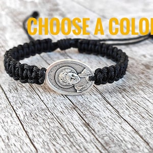 May include: Black braided bracelet with a silver oval charm featuring a depiction of Saint Carlo Acutis. The charm has the words "St. Carlo Acutis" inscribed on it.