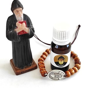 Op de afbeelding: Een beeldje van St. Charbel in een zwarte jurk met een rood boek, een kleine bruine fles met een wit etiket en een houten kralenarmband met een zilveren bedel. Op het etiket van de fles staat "CHARBEL".