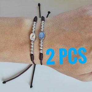 Könnte beinhalten: Zwei verstellbare Armbänder aus braunem Kordel mit silbernen Perlen und kleinen ovalen Anhängern. Ein Anhänger zeigt ein Vogelbild, der andere ein religiöses Bild. Der Text "2 PCS" ist blau.