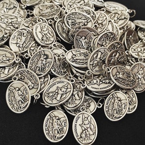 May include: A pile of silver oval-shaped charms with religious imagery. The charms feature images of angels, saints, and religious text. The charms are all made of metal and have a shiny finish.