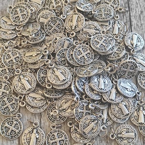 Puede incluir: Un conjunto de medallas de San Benito de color plateado. Cada medalla redonda presenta imágenes religiosas detalladas, incluyendo una cruz, figuras e inscripciones. Las medallas son pequeñas, con un lazo para sujetarlas a una cadena u otro artículo.