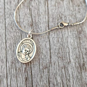 May include: A silver chain necklace with a silver pendant featuring a depiction of St. Carlo Acutis. The pendant is oval shaped and has a raised image of St. Carlo Acutis.
