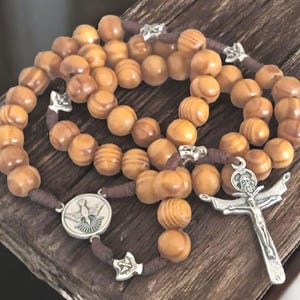 May include: A rosary with light brown wooden beads and a dark brown cord. The rosary features a silver crucifix and a circular pendant with a dove. The beads are arranged on a wooden surface.