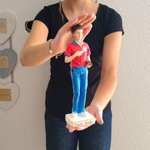 Op de afbeelding: Een beeldje van een jonge man in een rood shirt, blauwe jeans en witte schoenen, met een camera. Het beeldje staat op een witte basis met de naam "Carlo Acutis". De figuur wordt vastgehouden door een persoon in een zwart shirt.