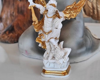 May include: White figurine of an angel with gold wings, standing on a gold base, holding a sword and defeating a demon.