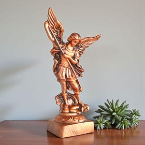 Op de afbeelding: Een bronzen beeld van een engel met uitgespreide vleugels, die een speer vasthoudt en boven een verslagen figuur staat. Het beeld staat op een vierkante basis en heeft ingewikkelde details. Rechts staat een kleine vetplant.