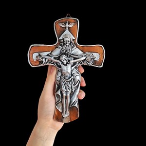 Może przedstawiać: Srebrny krzyż religijny z brązową drewnianą ramą. Krzyż przedstawia wizerunek Jezusa Chrystusa na krzyżu, z Bogiem nad nim i gołębiem na górze. Krzyż jest trzymany na czarnym tle.