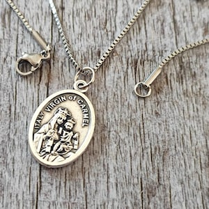 May include: Silver chain necklace with a silver oval pendant featuring a depiction of the Virgin Mary and baby Jesus. The pendant has the text "Italy Virgin of Carmel" inscribed around the edge.