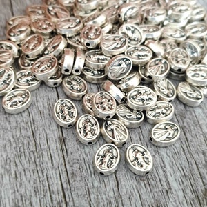 May include: A collection of silver-colored oval beads. Each bead features a detailed relief design of a religious figure. The beads are scattered on a wooden surface, creating a textured and visually appealing arrangement.