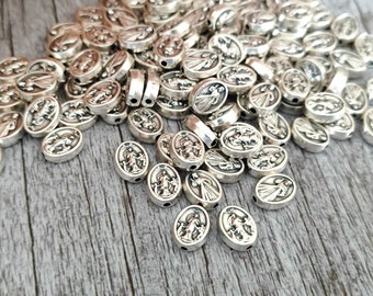 May include: A collection of silver-colored oval beads. Each bead features a detailed relief design of a religious figure. The beads are scattered on a wooden surface, creating a textured and visually appealing arrangement.