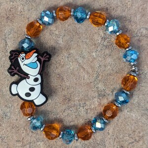 May include: A beaded bracelet featuring orange and blue faceted beads, interspersed with silver-toned spacers. The bracelet includes a charm of the character Olaf from the movie Frozen. The charm is white, black, and orange.