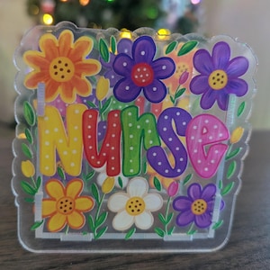 May include: A clear acrylic sign featuring the word Nurse in colorful, polka-dotted letters. The design is surrounded by vibrant illustrations of orange, purple, yellow, and white flowers with green stems and leaves on a transparent background.