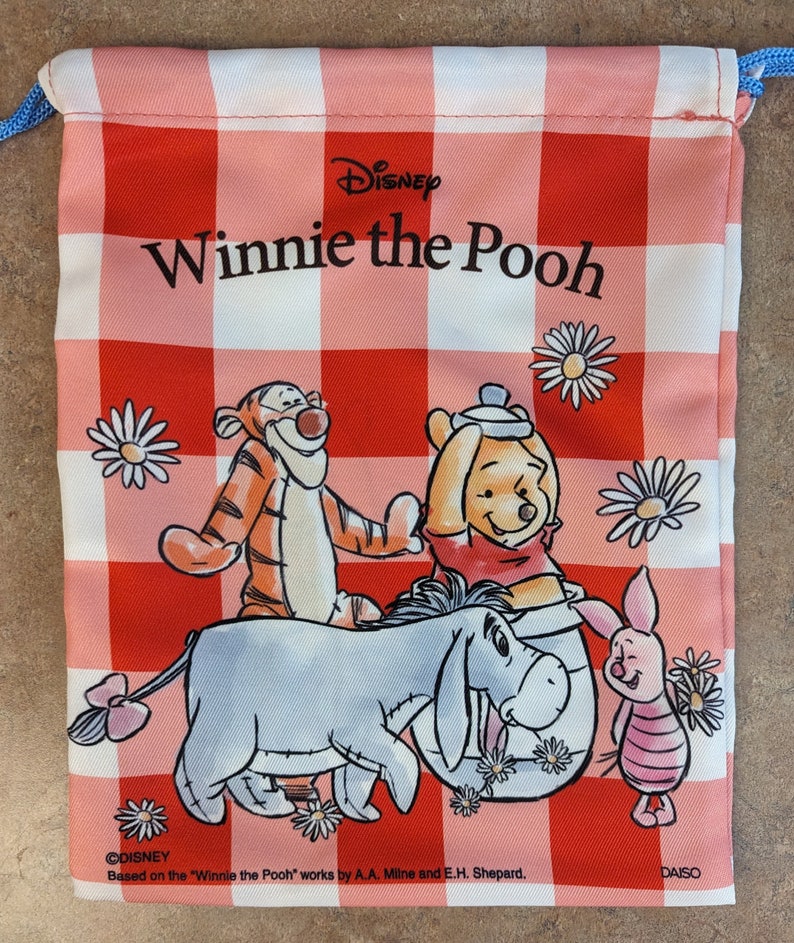 May include: A red and white checkered drawstring bag featuring Winnie the Pooh and his friends. The bag has the text "Disney Winnie the Pooh" and "&copy;DISNEY Based on the "Winnie the Pooh" works by A.A. Milne and E.H. Shepard. DAISO"