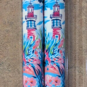May include: Two cylindrical objects with a repeating pattern of a lighthouse, flowers, and foliage. The lighthouse is red and white with a blue roof. The background features blue sky and white clouds. The floral elements are pink and the foliage is green.