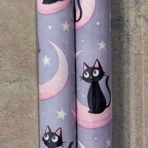 May include: Two cylindrical objects with a repeating pattern of black cats sitting on pink crescent moons against a lavender background. White stars are scattered around the moons. The cats have pink ears and large eyes.