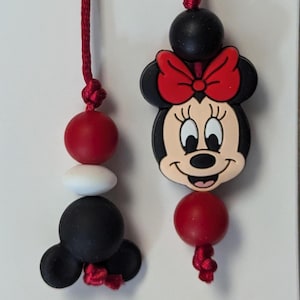 May include: A red, white, and black silicone necklace with a Minnie Mouse charm. The necklace has a red string and a black silicone bead with Mickey Mouse ears.