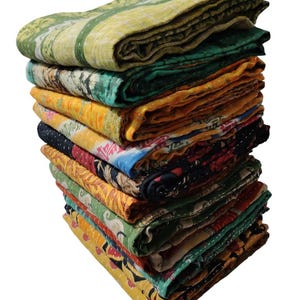 May include: A pile of folded, patterned quilts in various colours. The quilts display a range of designs, including floral and geometric patterns, in shades of green, yellow, and orange. The quilts are stacked, revealing their different textures.