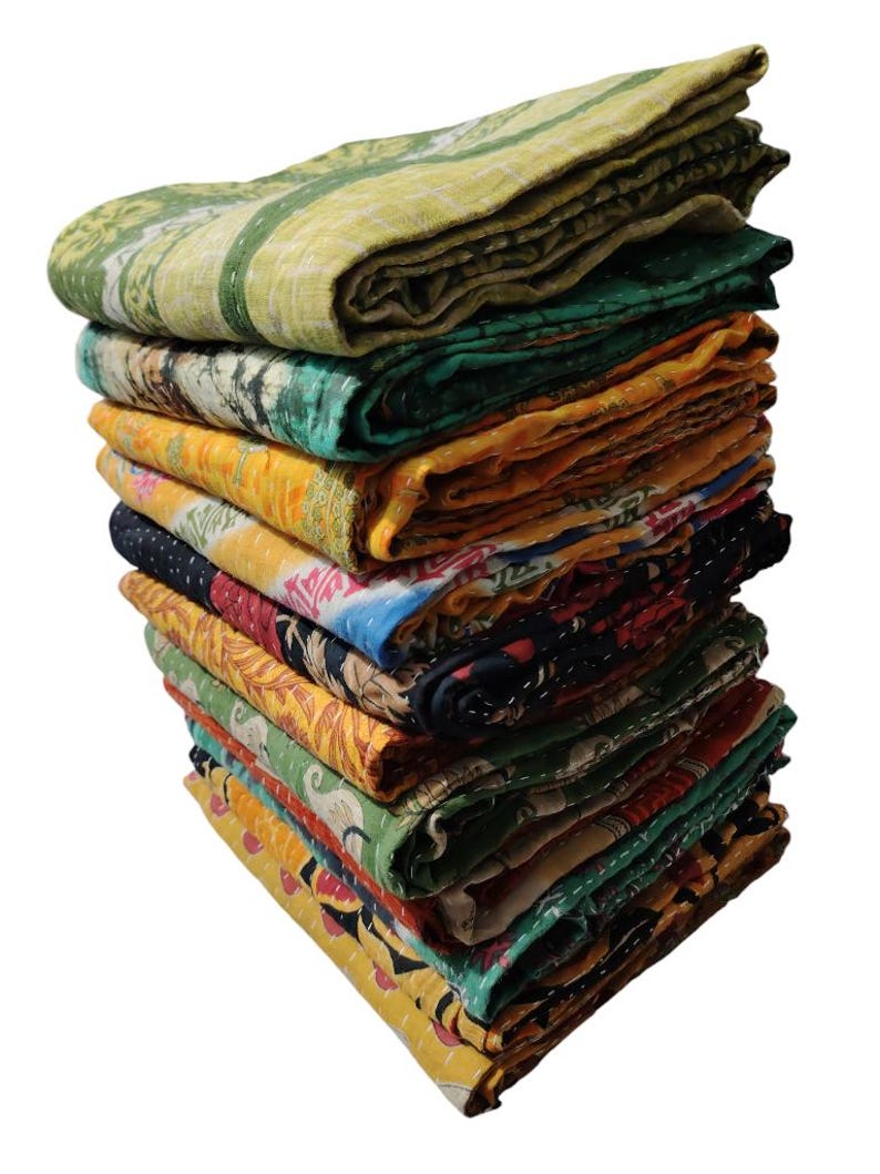 May include: A pile of folded, patterned quilts in various colours. The quilts display a range of designs, including floral and geometric patterns, in shades of green, yellow, and orange. The quilts are stacked, revealing their different textures.