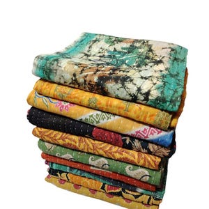 May include: A stack of colourful, patterned quilts. The top quilt has a teal and brown tie-dye design. Other quilts feature yellow, black, red, and green floral and geometric patterns. Each quilt is stitched with a contrasting thread.