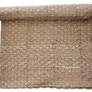 Op de afbeelding: Een opgerolde, bruin gekleurde quilt met een herhalend patroon van witte en donkergrijze bloemvormen. De quilt is met witte draad met de hand gestikt en de randen zijn geschulpt.