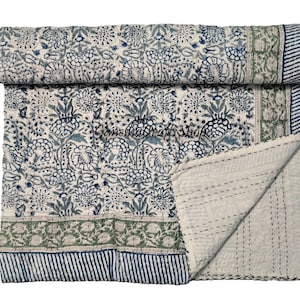 May include: A folded cotton quilt featuring a white base with a repeating blue floral block print pattern. The quilt includes a green floral border and a blue and white striped edge, finished with visible running stitch quilting details.