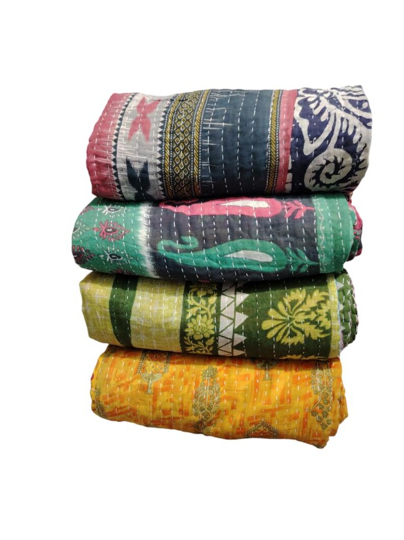 May include: A stack of four folded quilts in various colours. The top quilt features pink, grey, and navy blue with floral and geometric designs. The second quilt is green and pink. The third quilt is yellow and green. The bottom quilt is yellow.