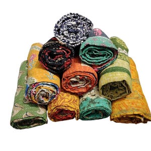 May include: A selection of rolled kantha quilts in various colours. The quilts feature diverse patterns and designs, such as floral, geometric, and abstract. The colour range includes yellow, orange, green, blue, and black.