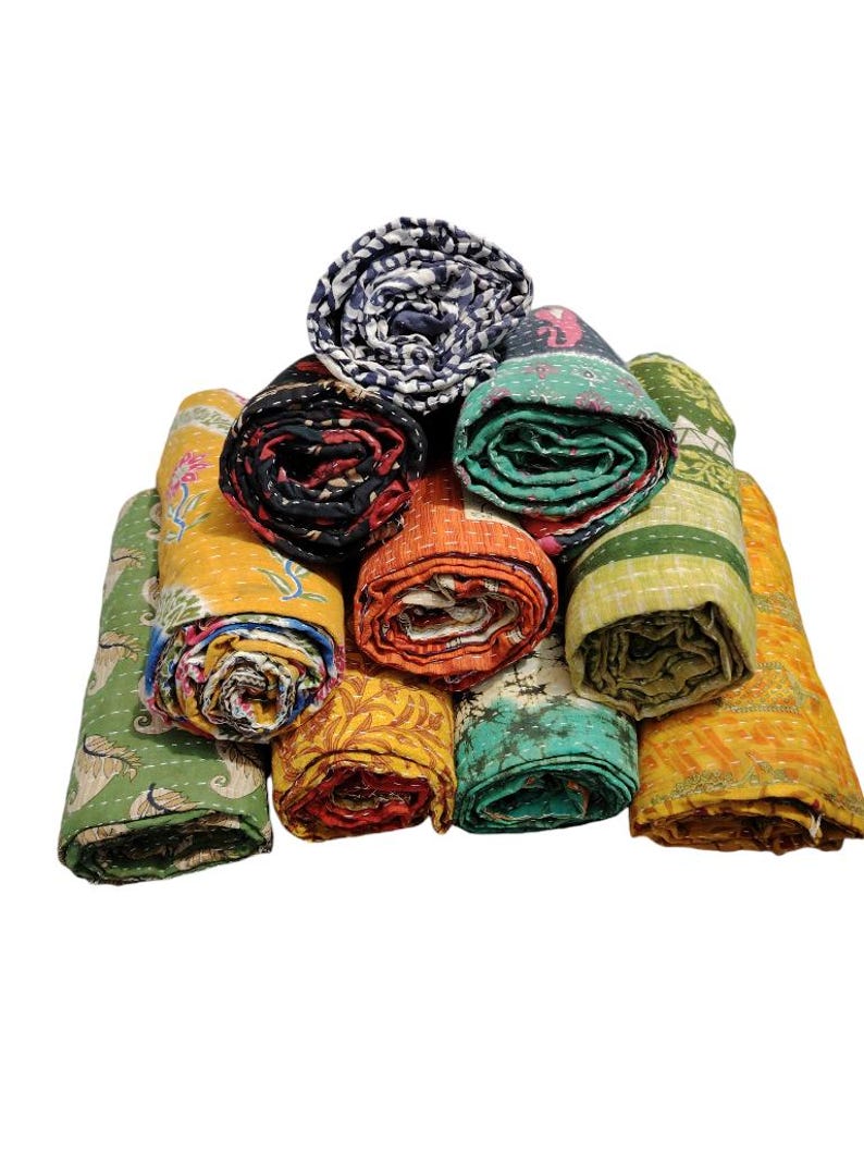 May include: A selection of rolled kantha quilts in various colours. The quilts feature diverse patterns and designs, such as floral, geometric, and abstract. The colour range includes yellow, orange, green, blue, and black.