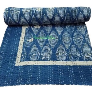 Op de afbeelding: Een indigo blauwe quilt met een wit geometrisch patroon, opgerold aan één uiteinde. Het uitgevouwen gedeelte toont hetzelfde ontwerp en een witte rand. De quilt is met de hand gestikt.