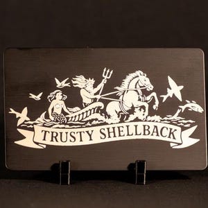 May include: A black rectangular plaque with a white illustration of a mythological scene. The image features a mermaid, a figure with a trident, a horse-drawn chariot, birds, and the text "TRUSTY SHELLBACK".