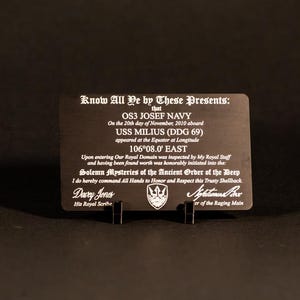 May include: A black metal plaque with white engraved text, held upright by a black stand. The text includes the words "Know All Ye by These Presents" and details about OS3 Josef Navy and the USS Milius (DDG 69).