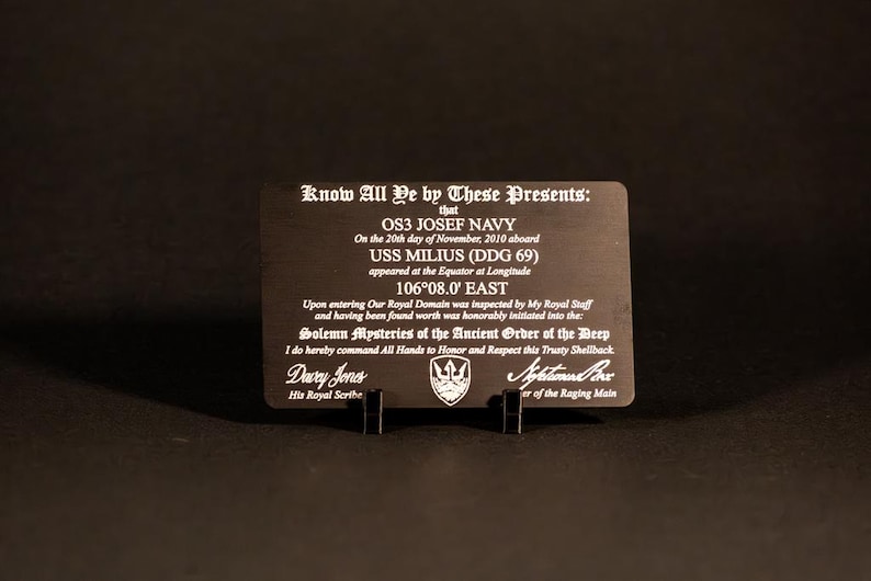 May include: A black metal plaque with white engraved text, held upright by a black stand. The text includes the words "Know All Ye by These Presents" and details about OS3 Josef Navy and the USS Milius (DDG 69).