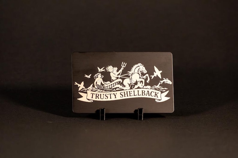 May include: A black rectangular plaque with a white illustration of a mythological scene. The image features a mermaid, a figure with a trident, a horse-drawn chariot, birds, and the text "TRUSTY SHELLBACK".