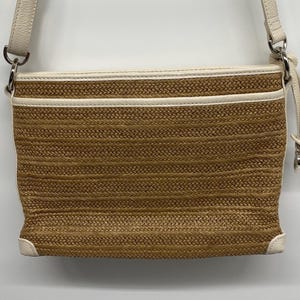 May include: A rectangular crossbody bag featuring a woven tan straw body with cream-colored leather trim. The bag includes a top zipper closure with a leather pull, silver-tone metal hardware, and an adjustable cream leather shoulder strap.