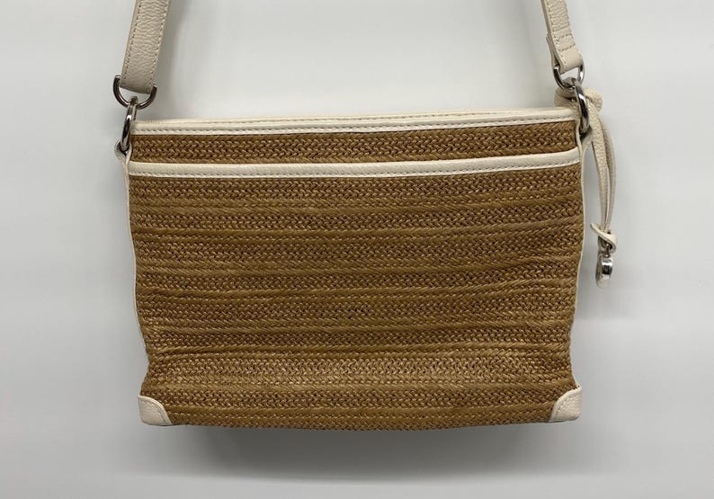 May include: A rectangular crossbody bag featuring a woven tan straw body with cream-colored leather trim. The bag includes a top zipper closure with a leather pull, silver-tone metal hardware, and an adjustable cream leather shoulder strap.