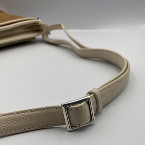 May include: A cream-colored shoulder bag strap made of textured leather with a silver-toned metal square buckle for length adjustment. The strap is attached to a tan-colored bag body that is partially visible in the upper left corner of the frame.